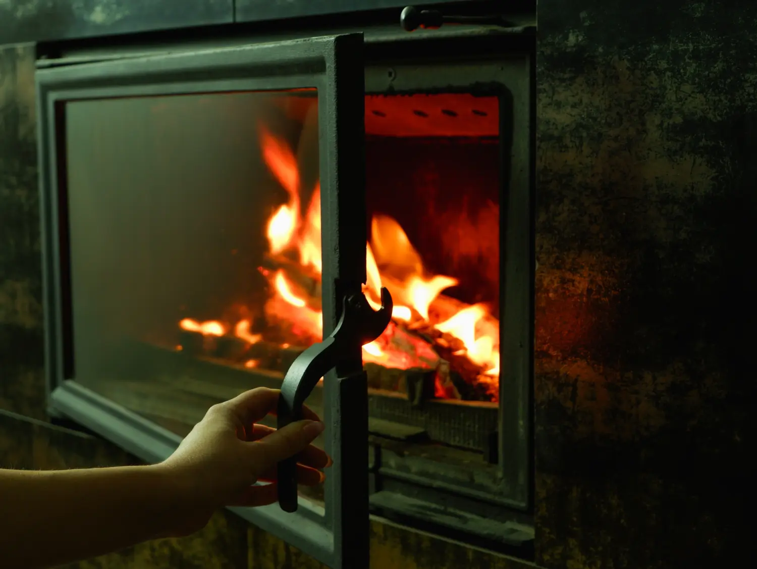 Die Hand einer Frau öffnet die Glastür des Kamins, in dem das Holz brennt